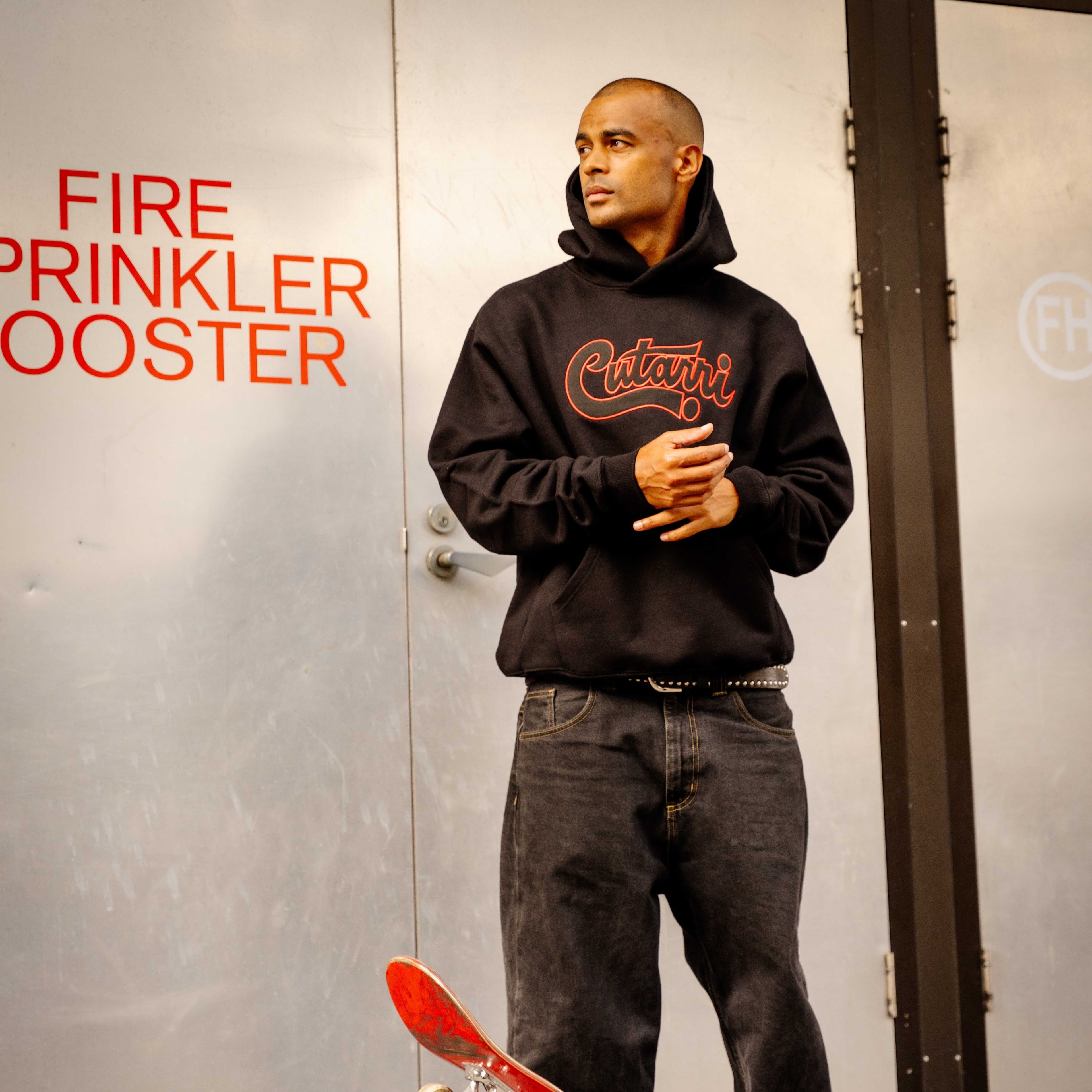 Man in a black hoodie holding a red skateboard against a metallic wall with text.