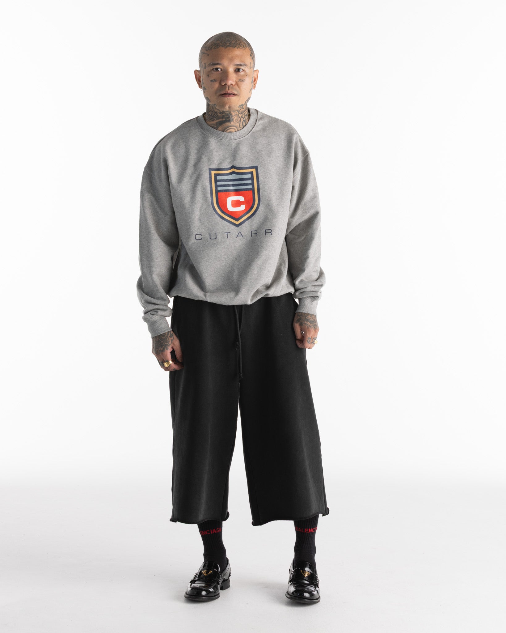 Person wearing a gray sweatshirt with a logo and text, black pants, and black shoes on a white background