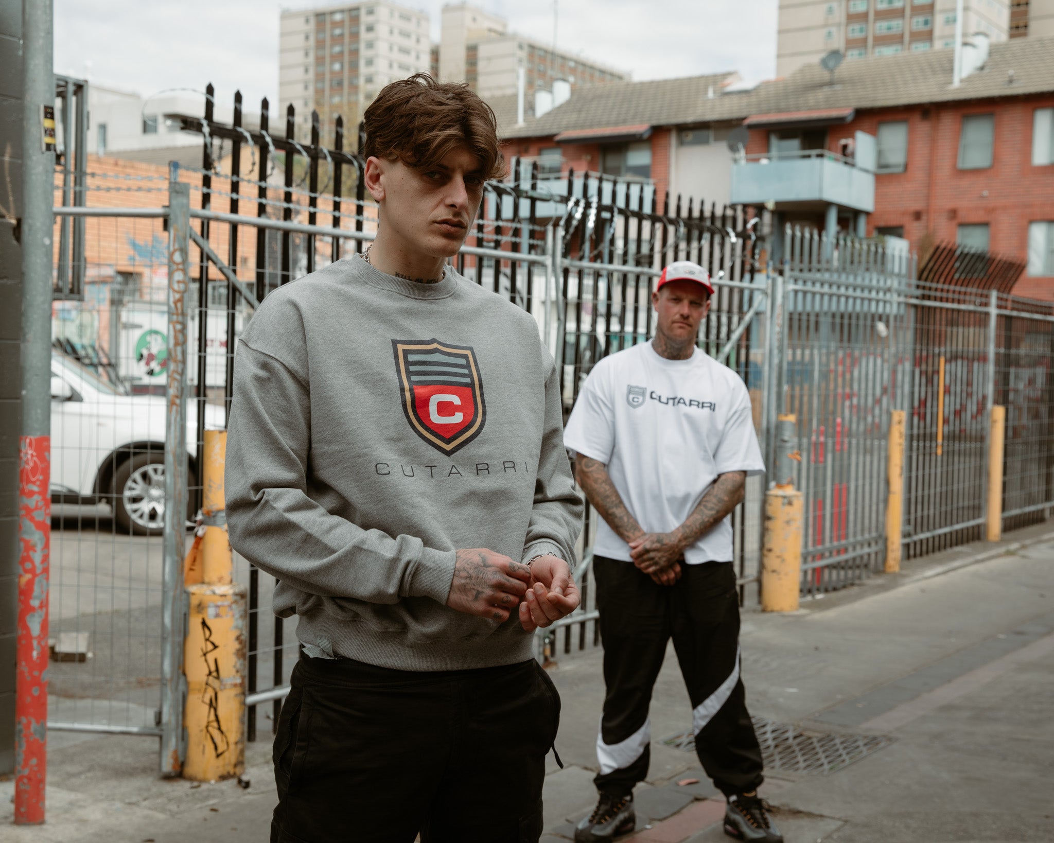 Two men standing on a street with a man wearing a gray sweatshirt with a logo.