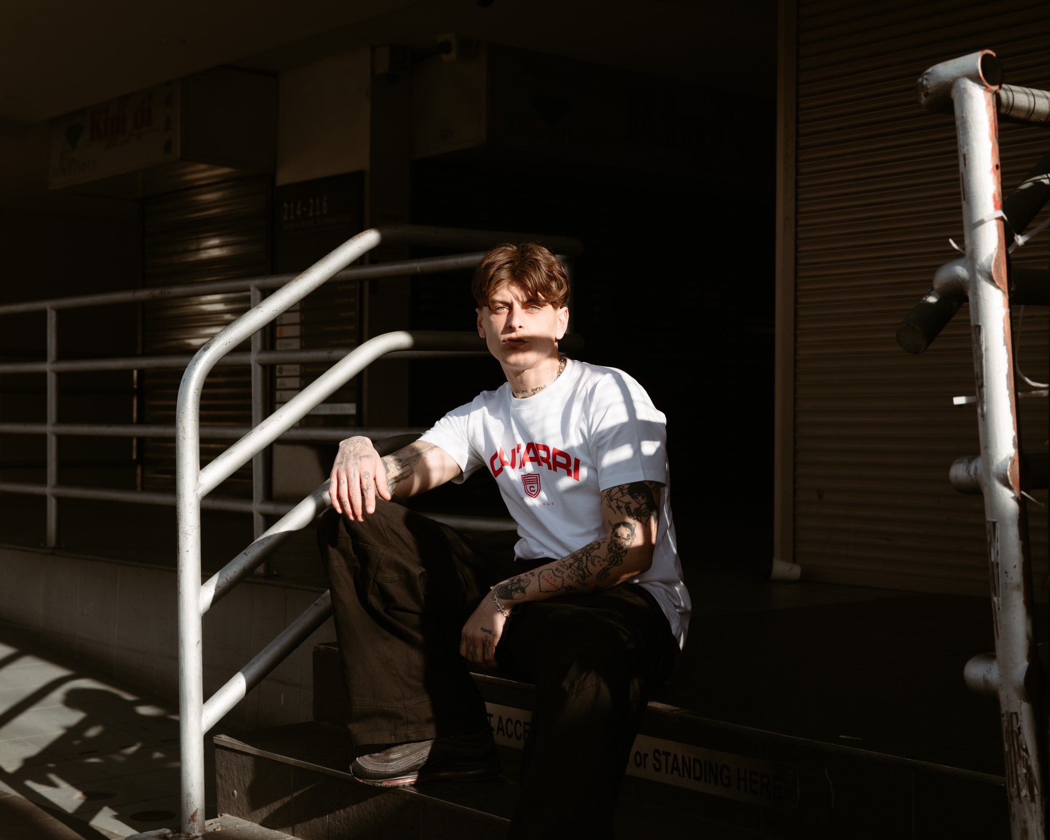 Person sitting on a staircase wearing a white t-shirt with red text, surrounded by industrial elements.