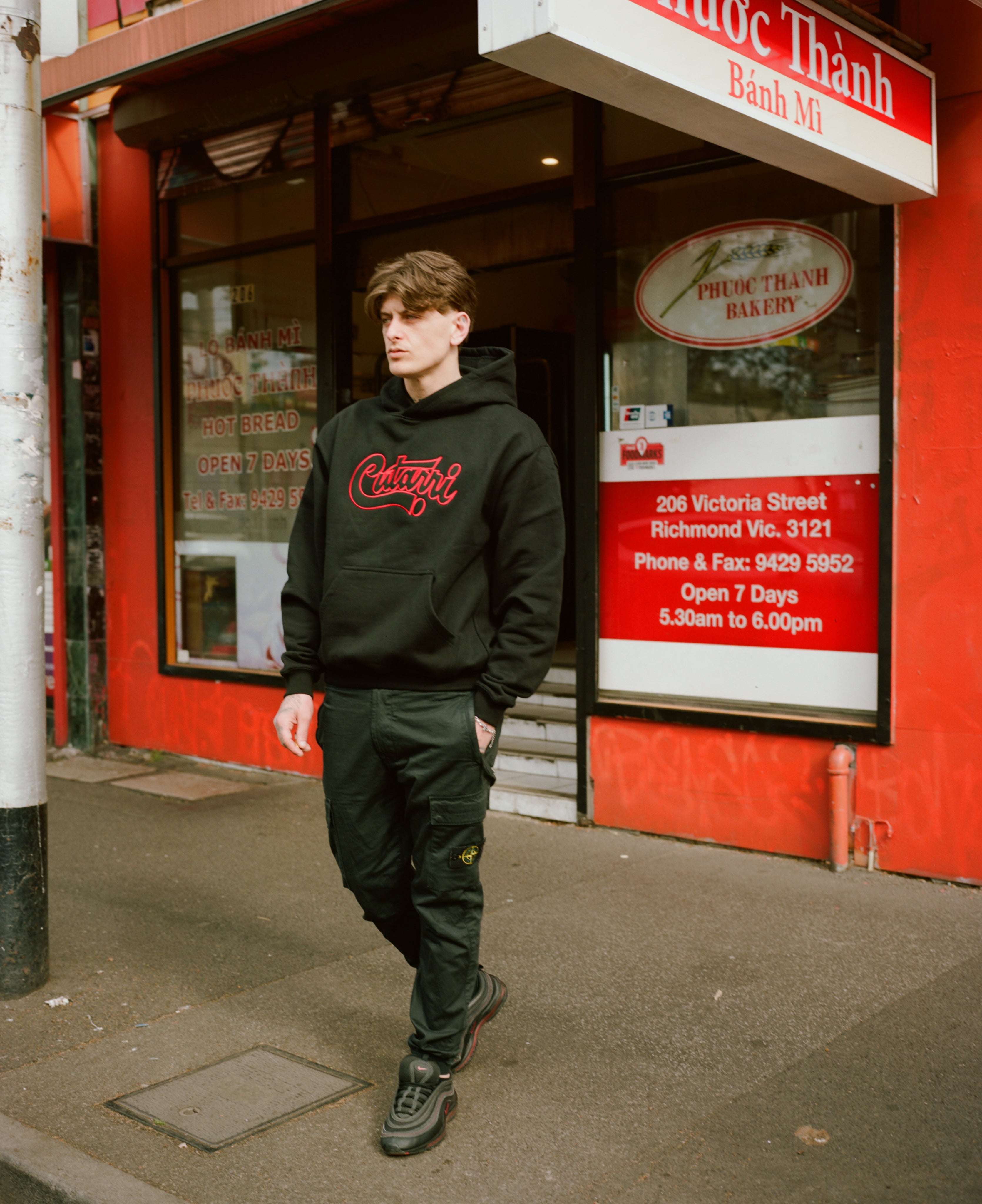 Person wearing a black hoodie with red text standing in front of a bakery storefront.
