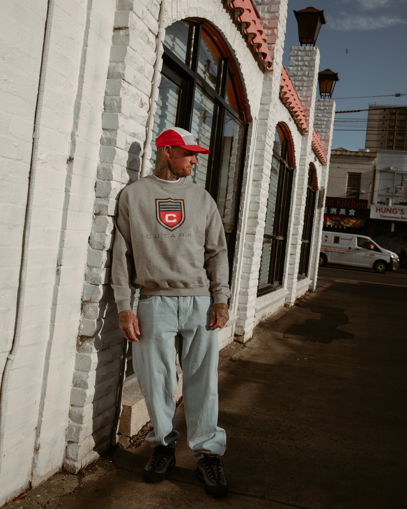Man wearing a gray sweatshirt with a logo and light blue pants standing against a white brick wall.
